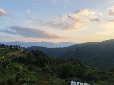 The image shows a vast mountainous landscape in the village of Leshten, Bulgaria, at dusk. The sky is a gradient of pale blue and warm yellow, with scattered clouds tinged with pink and orange. Layers of blue mountains recede into the distance. In the foreground, a steep, green hillside is dotted with trees and vegetation. A small cluster of rustic, stone-and-wood houses with corrugated roofs sits on the crest of the hill. A dirt road winds past the houses, with a dark-colored vehicle parked on it. The overall impression is one of a remote, tranquil rural setting bathed in the soft light of sunset.