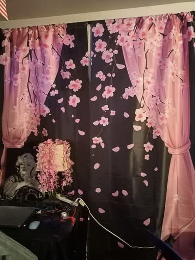 The image depicts an indoor scene featuring a black curtain with a cherry blossom pattern in shades of pink. The curtain is draped and tied back on the right side. To the left of the curtain, there is a black table with various objects, including a laptop, a computer mouse, cables, and a small container. Behind the table, a black and white portrait of Marilyn Monroe is visible. A lampshade adorned with pink flowers sits on the table. The overall scene suggests a personal living space. There is no information in the image to determine the city of Amarillo, United States of America, or any specific location details beyond it being an indoor environment.