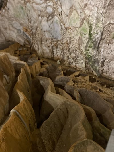 The image shows the interior of a cave, likely within the Zlatibor region of Cajetina Municipality, Serbia. The cave walls are rough and textured, composed of layered rock in shades of white, grey, and brown, with some patches of green algae or moss. In the foreground, the floor of the cave is covered with uneven, eroded rock formations that create a series of undulating ridges and hollows. These formations appear to be made of a lighter-colored, sandy rock, with some areas showing a yellowish or orange hue. The lighting suggests a natural cave environment, with shadows accentuating the contours of the rock. There are no people or artificial objects visible in the frame.
