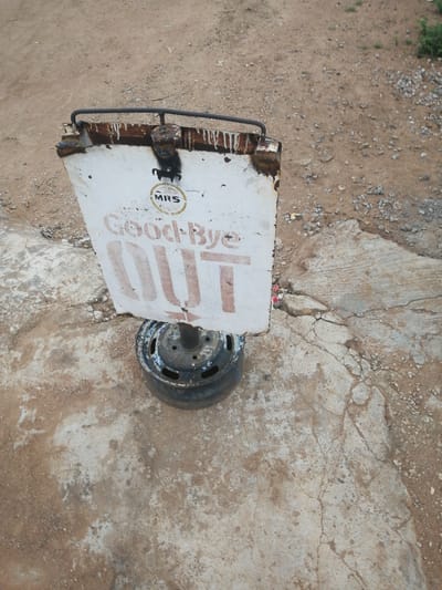 A white sign with "Good-Bye OUT" written in red letters stands on a dirt and concrete surface in Ajasse Ipo, Nigeria. The sign is weathered and rusted at the top. A black silhouette of a horse or similar animal is visible above the text. The sign is mounted on a pole that is attached to an old, rusty car wheel, serving as a base. The surrounding area is a mixture of loose dirt, gravel, and cracked concrete. The lighting suggests it is daytime.