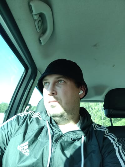 A man in his 30s, with light skin, green eyes, and short brown hair, wears a black bucket hat, a white t-shirt, and a black Adidas jacket with white stripes. He is in the passenger seat of a car, looking out the open window at a rural landscape. The car's interior is visible, including a ceiling handle, and the sky is blue and clear. The image was taken in Drahňovice, Czechia, on a sunny day.