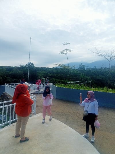 The image captures an outdoor scene at a viewing platform overlooking a lush, hilly landscape, likely in Pangalengan, Indonesia, known for its tea plantations and scenic views.
In the foreground, three individuals are prominent on a concrete platform. On the left, a woman wearing an orange hijab and top, with light-colored pants, stands partially turned away, observing the scene. Next to her, a young girl in a black hijab, light pink top, and pink pants stands facing forward, looking down at something in her hands. To the right, another woman, smiling broadly, wears a light purple hijab, a white long-sleeved shirt, black pants, and white sneakers; she holds a smartphone up, seemingly taking a photo or video of the panoramic view or the people in the background.
Behind these individuals, a white metal railing separates the foreground platform from a lower section where several other people, mostly women also wearing hijabs, are gathered. Some in the background are also holding phones, actively engaging with the scenery. A faint "10" is visible on an object or clothing in the mid-ground.
The background features extensive green hills and mountains under an overcast sky with scattered clouds, indicating a cloudy but bright daytime. A tall, thin pole stands on the left side of the frame, and a few slender trees with sparse foliage dot the landscape, including one prominent tree branch on the right. The overall atmosphere suggests a leisurely outing, with people enjoying the natural beauty and documenting their experience.