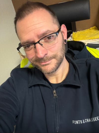 A male individual is depicted in a close-up, self-taken photograph within an indoor setting in Grevenmacher, Luxembourg. The man, appearing to be in his 30s to 40s, has short brown hair, a short beard, and is wearing black-framed eyeglasses. A black ear gauge is present in his left ear. He is dressed in a dark navy blue zip-up jacket with the white embroidered text "PONTS & CHAUSSÉES" visible on the right chest area. A neon yellow high-visibility garment is observable over his left shoulder and upper back. The background elements include a light-colored wall to the left, and to the right, a dark object, a partially visible box labeled "BATTERY", and a yellow cloth or towel. The individual is looking directly at the camera with a slight smile.