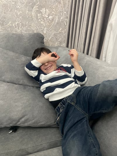 A child lies upside down on a gray couch. The child is wearing blue jeans and a striped long-sleeve shirt. The child is smiling and covering their eyes with one hand. The couch is in a room with gray wallpaper and curtains. The location is Akhalkalaki, Georgia.