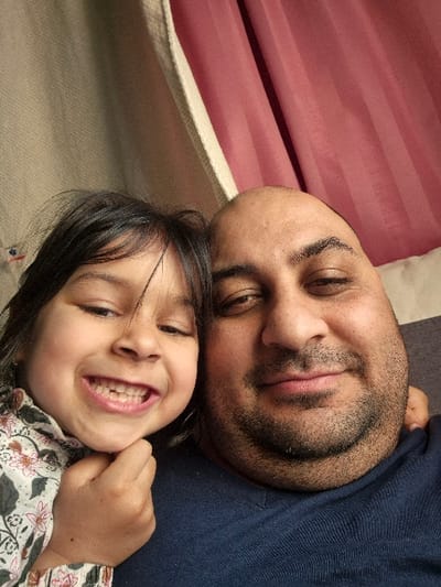 The image captures a tender and joyful moment between an adult male and a young child, likely a father and daughter, in what appears to be an indoor setting. The image is rotated 90 degrees clockwise from its natural orientation, showing the subjects lying down together.
In the foreground, taking up the majority of the frame, are the faces of the two individuals. The adult male, with dark, closely-cropped hair and a short, dark beard or stubble, is positioned on the lower half of the frame (when viewed as rotated). His eyes are partially closed or looking softly downward, and a gentle, relaxed smile plays on his lips, conveying a sense of peace and contentment. He is wearing a dark blue, possibly knitted or textured, top or sweater.
Resting closely beside him, and slightly above (when viewed as rotated), is the young child. She has dark hair and a bright, infectious, wide-open smile, showing her teeth. There appears to be a gap from a missing front tooth, which is common in young children and adds to her charming expression. Her eyes are wide open and sparkling with joy. Her left hand is visible, curled under her chin or cheek, contributing to her playful pose. She is wearing a light-colored top adorned with a floral pattern featuring dark green leaves and pink or reddish flowers.
The two individuals are positioned very close together, with the child's head resting on or very near the adult's head/shoulder area, emphasizing their affectionate bond. The overall mood is one of warmth, happiness, and closeness.
The background is softly blurred. To the upper left (when viewed as rotated), behind the child's head, there is a light beige or off-white fabric, possibly a blanket or sheet. To the right, a reddish-pink fabric with visible horizontal folds, possibly a curtain or part of a bedspread, fills the rest of the background. The lighting is soft and even, suggesting an indoor environment, perhaps during the day or evening. No text is visible in the image.