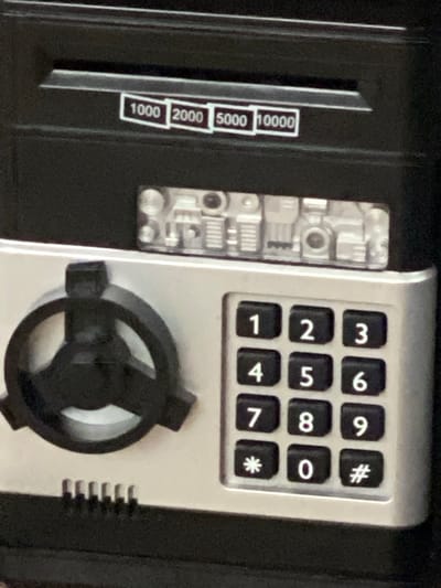 The image shows a close-up of a black and silver electronic safe. On the top black section, there is a slot with white labels above it that read "1000", "2000", "5000", and "10000". Below this slot is a clear plastic panel with visible electronic components and what appears to be a small speaker. The lower silver section features a rotary dial with three prongs on the left and a numeric keypad on the right. The keypad has buttons labeled 0 through 9, as well as "*" and "#". The safe appears to be sitting on a dark surface, possibly a table or shelf. There is no indication of the safe's location in Ispas, Ukraine.