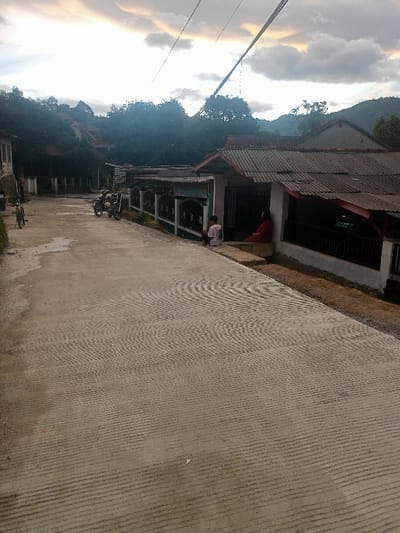 This image presents a serene, semi-rural street scene in Kecamatan Pangalengan, Indonesia, likely captured during the late afternoon or an overcast day. The focal point is a concrete road, characterized by distinctive patterned grooves – both parallel lines and areas with ripple-like textures – which extend from the foreground towards the background, gently curving to the left.
On the right side of the road, a row of houses is visible. The closest house features a dark, corrugated metal roof and a light-colored base. Two individuals are seated on a low step or porch in front of this residence. One person, positioned closer to the viewer, wears a white shirt with a dark emblem on the back. The other, seated slightly behind and to the right, is dressed in a red garment, possibly a long dress or robe. A dark, ornate fence separates the house property from the roadside.
To the left of the road, two motorcycles are parked, and further down, a bicycle is also visible. The background is dominated by lush green trees and dense foliage, covering gentle hills that recede into the distance. Fainter silhouettes of higher mountains or hills can be seen beyond.
The sky is largely overcast, providing diffused light, with lighter patches and subtle hints of pale orange or pink, suggesting either an impending sunset or a soft, cloudy daylight. Utility lines traverse the upper section of the image. The overall impression is one of tranquility and everyday life in a quiet village setting. No visible text is present in the image.