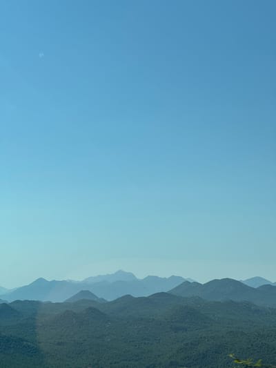 A landscape photograph captures a series of rolling, verdant hills and mountains under a clear, pale blue sky. The foreground is dominated by dense, dark green foliage covering the undulating terrain. As the eye moves towards the background, successive layers of mountains are visible, their forms softened and rendered in progressively lighter shades of blue and purple due to atmospheric perspective. The farthest peaks are faint silhouettes against the horizon. The sky is vast and unblemished, with a slight gradient from a lighter hue near the horizon to a deeper blue overhead. There are no people or man-made structures visible in the scene. The image appears to be taken from an elevated vantage point, overlooking a vast natural expanse. The location is identified as Dobrsko selo, Montenegro.