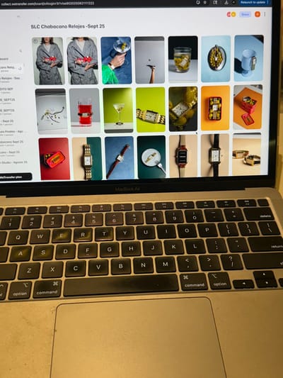 An Apple MacBook Air laptop, silver in color, is positioned at a slight downward angle, its screen illuminated and displaying a web browser. The browser, indicated by `wetransfer.com` in the URL bar, presents a grid of 20 thumbnail images under the title "SLC Chabacano Relojes -Sept 25." These thumbnails feature various product photographs, predominantly showcasing wristwatches. Specific compositions include watches worn by individuals, integrated into cocktail glasses with olives, displayed on forks, or packaged in small tins. The laptop's keyboard shows signs of wear, with keys such as Q, W, E, R, T, Y, A, S, D, F, and G exhibiting a yellowish discoloration. The trackpad is visible below the keyboard. The laptop rests on an unidentifiable flat surface, with the background out of focus and dimly lit. No individuals are visible interacting with the laptop. This scene is located in San José del Cabo, Mexico.
