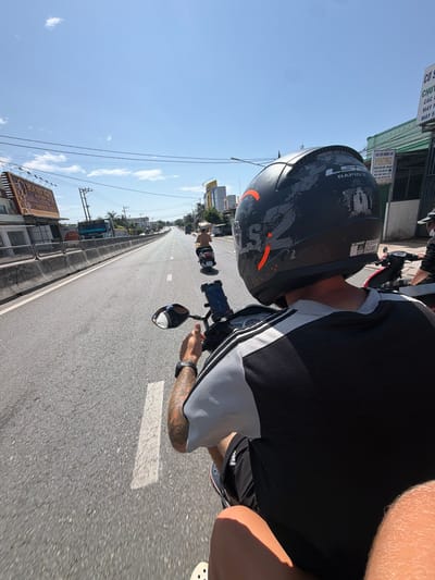 This media file captures a first-person perspective, rotated 90 degrees clockwise, of someone riding a motorcycle on a clear, sunny day in what appears to be Cam Ranh Ward, Vietnam.
**Subjects:** The primary subject is the rider or passenger, seen from the waist up, wearing a black and white long-sleeved shirt and a black helmet. The helmet has an orange graphic design and text "RAPID II" and "LS2" visible on it. Tattoos are visible on the person's right forearm, which is gripping the motorcycle's handle. Another person on a motorcycle is partially visible in the distance on the left side of the road.
**Objects:** The motorcycle's handlebars, a side mirror, and a phone mounted on the handlebars are clearly visible. The asphalt road stretches ahead, with white lane markings. Buildings line both sides of the road, featuring various signs and architectural styles common to the region. Overhead power lines crisscross the bright blue sky, which has a few scattered clouds.
**Scene/Setting:** The scene depicts a busy daytime street, likely a main road or highway given its width and the type of buildings alongside it. The setting is urban or semi-urban.
**Activity or event taking place:** The image captures the experience of riding a motorcycle, suggesting travel or commuting.
**Notable details:** The weather is bright and sunny with a clear blue sky, indicating daytime. The strong sunlight casts shadows. The environment is typical of Southeast Asian streets, with visible power lines, local signage, and common modes of transport. The image's orientation is sideways.
**Visible text:**
* "HOA SEN HOM THÉP" (a building/business sign)
* "CƠ SỞ ĐIỆN CƠ PHONG" (another business sign, likely an electrical appliance/motor shop)
* "CÁC LOẠI MOTOR"
* "MÁY PHÁT ĐIỆN"
* "RAPID II" (on the helmet)
* "LS2" (on the helmet)
* "2985 559 437" (phone number)
* Partially visible Vietnamese text like "COS," "CHU," "CÁC," "MÁY F," and "MÁY S".