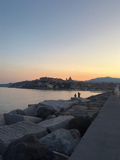 The image depicts a coastal scene at dusk in Imperia, Italy. The sky displays a gradient of colors, transitioning from a pale yellow at the horizon to a soft blue overhead, indicating sunset. The water reflects the fading light.
In the foreground, a man-made jetty or seawall is constructed of large, rough-hewn rocks. Two silhouetted figures are visible on the rocks near the water's edge, appearing to be interacting or observing the surroundings. Further along the jetty, a paved walkway runs parallel to the rocky structure.
Beyond the water, a town is nestled against a backdrop of hills. The buildings of the town are illuminated by lights, suggesting that evening has fallen. The lights of the town twinkle against the darkening landscape. The overall atmosphere appears serene and peaceful, capturing the tranquil beauty of the Italian coast at twilight. No visible text is present in the image.