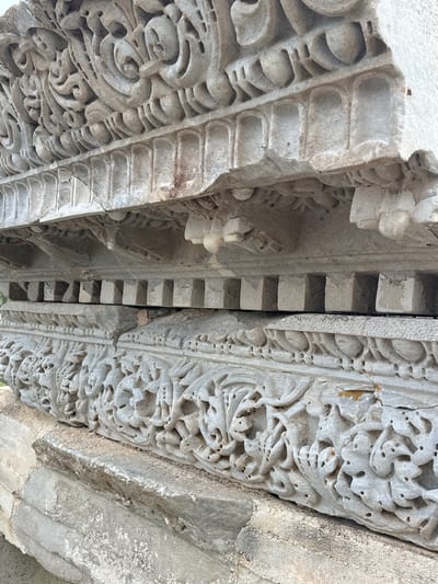 This close-up image captures an intricate section of carved stone architecture, likely part of an ancient or historical structure. The primary subjects are the detailed carvings themselves, with no people visible. The scene appears to be an exterior or partially enclosed space, bathed in bright, natural daylight, suggesting it is daytime.
The image shows three distinct vertical sections of carved stone. On the left, a panel features a repetitive pattern of interconnected, rounded, and elongated shapes, possibly stylized leaves or scrolls, with small rectangular indentations. The central section consists of a series of raised, rectangular blocks arranged in a stepped, interlocking manner, forming a deep recess or joint between the two larger carved elements. The rightmost panel showcases highly ornate and dense carvings, characterized by swirling, organic forms, intricate vegetal or floral motifs, and numerous small holes or depressions that add to their three-dimensional quality.
The stone itself is light-colored, possibly limestone or marble, and shows signs of significant age and weathering. There are visible cracks, chips, and areas of discoloration, including brownish-yellowish stains, hinting at prolonged exposure to the elements. The texture of the stone appears somewhat coarse. The perspective is angled upwards, emphasizing the depth and complexity of the overhead or vertical architectural decorations. No specific activity or event is taking place. There is no visible text. Given the context of Datça, Turkey, these carvings likely represent a style from a historical period prevalent in the region, such as Byzantine, Ottoman, or ancient Greek/Roman influences.