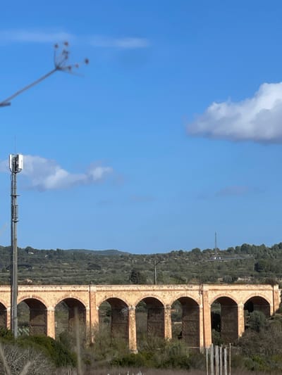 The image displays a daytime, panoramic view of a rural or semi-rural landscape in la Riera de Gaià, Spain, under a clear blue sky with scattered white cumulus clouds. In the mid-ground, a multi-arched, light-colored stone viaduct or aqueduct spans horizontally across the frame. To the left of the viaduct, a tall, metallic cellular communication tower with multiple antenna panels is prominently positioned. The background consists of rolling hills extensively covered with olive groves and other green vegetation. Another, thinner communication or utility tower is visible on a distant hill to the right. Overhead lines traverse the landscape above the viaduct. In the immediate upper-left foreground, an out-of-focus plant stem with small, dark seed heads extends into the frame. In the lower right foreground, several vertical, dark wooden or metal posts are visible amidst sparse ground cover. No human figures are present.