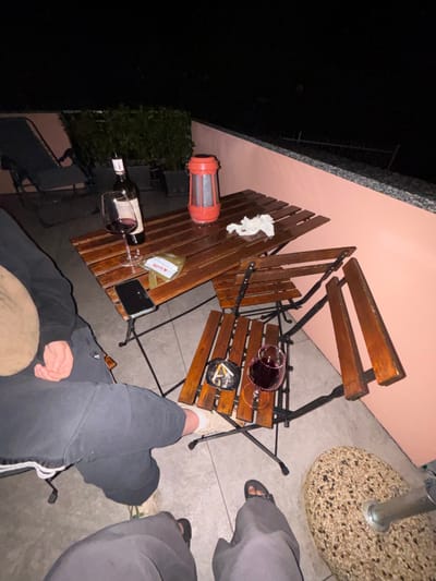 This image depicts an outdoor patio scene at night in Nesso, Italy. Two individuals are partially visible, seated or standing in the foreground. One person on the left wears dark athletic shorts or sweatpants with white trim, light-colored sneakers, and has a tan-colored object (possibly a hat or upper garment) near their head. A hand rests on their knee. The second person, at the bottom of the frame, wears dark pants and dark open-toed sandals.
The central focus is a wooden-slatted patio furniture set. A larger wooden table holds an uncorked bottle of red wine, a wine glass containing red wine, a black smartphone placed face-down, a small transparent packet labeled "MARACU," a crumpled white tissue, and a red portable lantern with a textured base. To the right, a smaller wooden-slatted chair serves as a surface, holding another wine glass with red wine and a dark circular ashtray containing multiple cigarette butts and ash. A third wooden chair, folded or partially collapsed, is positioned behind the smaller chair.
In the background, to the left, a dark grey lounge chair is visible, positioned near potted green foliage. A low, peach-colored concrete wall with a darker, speckled top edge runs along the right side of the patio. Beyond this wall, a dark, unlit area and a faint metal railing are discernible, suggesting a drop-off or lower terrace. The ground is paved with light-colored tiles. The scene is illuminated by artificial light sources, including the red lantern, against an otherwise dark night sky.