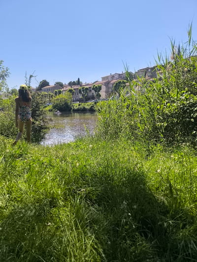 A medium shot depicts a sunny day with a young person in a swimsuit and yellow cap walking toward the Oignin river in Chézenas, France. The person is near the left edge of the frame, moving from left to right. The river reflects the clear blue sky. A row of houses with red tile roofs sits on the opposite bank. Lush green vegetation fills the foreground and right side of the frame. A small boat is visible on the river. A church spire appears behind the vegetation on the right.