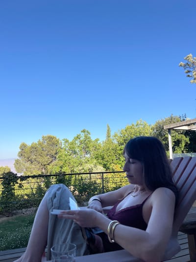 An adult female is depicted seated outdoors in a wooden slatted lounge chair within the Merom HaGalil Regional Council, Israel. She has dark hair with bangs, wears a dark red camisole top, light-colored trousers, a necklace, a black wristband, and two gold-colored bangles on her right wrist. Her gaze is directed downwards, focused on a small, light-colored rectangular object held in her left hand, which rests above a dark fabric or bag on her lap. A clear glass containing liquid is partially visible on a surface in the lower left foreground. The background features a clear blue sky, extensive green trees and foliage, and a distant landscape visible on the far left horizon. A black metal railing separates the immediate foreground from the verdant background. A wooden pergola structure is partially visible in the upper right background, and the ground beyond the railing shows grass with small white flowers.