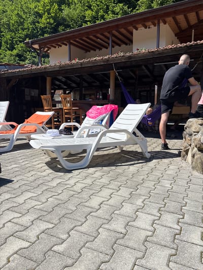 In Liava Reka, Bulgaria, a sunny outdoor scene unfolds, featuring a paved patio area with several white plastic lounge chairs. Two chairs in the foreground are empty, one with a white hat and black clothing placed on it. Another chair further back has an orange cushion. In the background, a covered patio structure with a wooden roof and tiled roof extends across the frame. Behind the structure, lush green trees form a dense forest. A person in a black t-shirt and shorts is partially visible on the right side of the image, leaning on a stone wall. A purple hammock is suspended between two posts under the covered area, with a person inside. Another person is visible in the background, sitting at a bar. Bottles are visible on a counter within the covered area. The overall setting appears to be a recreational or resort area.