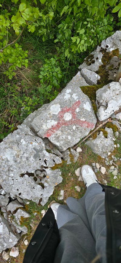 The image, likely taken at Kopito, Montenegro, depicts a section of a rough stone wall partially covered in moss and lichen, with surrounding vegetation. A red cross with a central white circle is painted on one of the stones. To the right, a person is standing with their foot on a stone near the wall. They are wearing grey sweatpants and white athletic shoes with black stripes. A portion of a black jacket or coat is also visible. The ground around the wall consists of scattered rocks, grass, and dry vegetation.