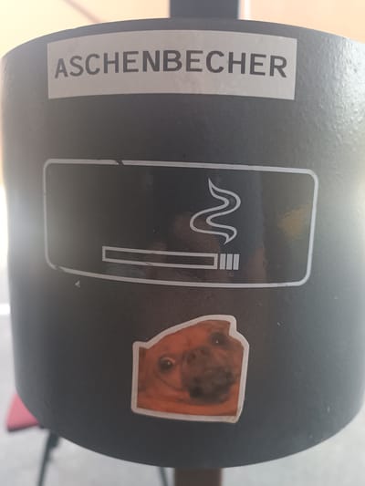 This media file is a close-up shot of a black ashtray, likely located in Bruchsal, Germany, given the visible German text. The ashtray is cylindrical and appears to be made of metal.
Prominently displayed on the ashtray is a white sticker with the German word "ASCHENBECHER," which translates to "ashtray." Below this text is a graphic depicting a cigarette with smoke rising from it, indicating the ashtray's intended use. The graphic is rendered in a stylized, outline form.
Adding a whimsical touch, a small sticker of a dog's face is affixed to the lower portion of the ashtray. The dog appears to be a fawn-colored breed, possibly a pug or a similar breed, with its eyes and nose clearly visible. The dog's expression is somewhat cartoonish, adding a lighthearted element to the otherwise functional object.
The background of the image is blurred, but it suggests an outdoor setting, possibly a public area such as a street, park, or cafe. The lighting indicates it is daytime, with a slight glare on the surface of the ashtray suggesting sunlight. The overall impression is a mundane object that has been personalized with stickers.