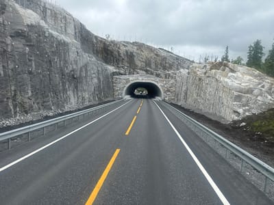 This image depicts a road leading into a tunnel, set against a backdrop of rugged, mountainous terrain. The road is paved and marked with yellow and white lines, with a silver guardrail running along either side. The tunnel entrance is reinforced with stone and concrete. The surrounding landscape is characterized by steep, rocky slopes, some of which are stabilized with netting and anchors. Trees and vegetation are visible on the higher slopes and in the distance. The sky is overcast, suggesting a cloudy or possibly rainy day. There are no people visible in the image, and the scene appears to be a natural landscape with significant infrastructure development. The location is identified as Grane, Norway. The overall impression is one of a modern road construction project in a remote, natural environment.