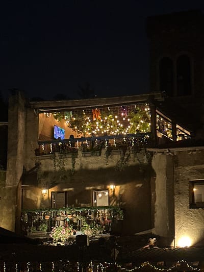 A building at night, illuminated with string lights, has vines growing on the side. Inside a balcony-like area, numerous lights twinkle, creating a festive atmosphere. A blue, rectangular object sits on the floor of this space. The building's exterior is rough and appears old, with several windows and a darkened upper section. Overall, the scene suggests a lively, outdoor setting in the dark.