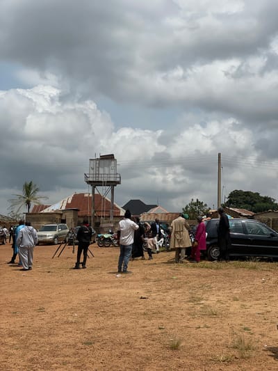 The image depicts an outdoor scene in Kassa, Nigeria, under a cloudy sky. The ground is covered in reddish-brown dirt with sparse patches of dry grass. In the background, several buildings with corrugated metal roofs are visible, along with a water tower on a metal frame. A palm tree stands to the left of the frame.

A group of people are gathered in the foreground and mid-ground. Some individuals are standing near parked vehicles, including a silver SUV, a black sedan, a motorcycle, and a three-wheeled vehicle. The people are dressed in various casual and traditional attire. Some are facing away from the camera, while others are conversing. One person is standing near a tripod. The overall atmosphere appears to be that of a community gathering or event.