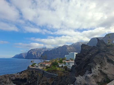This is a wide-angle, eye-level shot of the Los Gigantes cliffs on the island of Tenerife, Spain, in the municipality of Santiago del Teide. The image features the dramatic volcanic rock formations of the cliffs, which are a dark, rugged texture. The blue ocean stretches out to the left, meeting the base of the cliffs.
In the foreground on the right, a large, dark, rocky outcrop dominates the view. To the left of this outcrop, along the cliff edge, is a small development of white buildings, some with red-tiled roofs, set amidst lush green trees and palm trees. Further back, a large, multi-story white hotel with blue accents is visible, perched on the cliffside.
The sky above is a vibrant blue, with scattered white, fluffy clouds. The overall impression is one of a coastal resort area nestled against a majestic natural landscape. There are no people visible in the image. The scene appears to be captured during the daytime, with ample sunlight illuminating the landscape.
