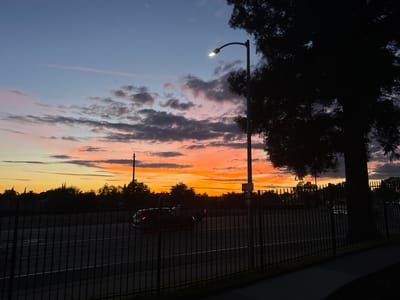 The image depicts a sunset scene in Sacramento, California. A streetlamp with its light on illuminates the foreground, casting a glow against the darkening sky. In the middle ground, a black SUV drives along a road, its taillights visible. A black metal fence runs across the lower portion of the image, separating the viewer from the road. The background features a silhouetted landscape of trees and buildings against the vibrant colors of the sunset. The sky transitions from a deep blue at the top to hues of orange, pink, and yellow near the horizon, with scattered clouds adding texture to the scene. The overall impression is one of a tranquil evening in an urban environment.