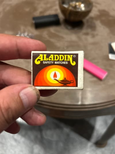 The media file shows a close-up of a person's hand holding a small box of Aladdin Safety Matches. The matchbox is predominantly brown with a graphic design featuring concentric circles of orange, yellow, and red, with a depiction of a lamp and a flame at the center. The brand name "ALADDIN" is written in a stylized yellow font, and "SAFETY MATCHES" is written below it in white text.
In the background, slightly out of focus, are other objects. To the left, there's a dark cylindrical object, possibly a battery or a pen. Further left and behind it, a metallic, round object with a textured surface is visible, which could be part of a lamp or another decorative item. A bright pink object, also out of focus, is positioned behind the matchbox, suggesting it might be a marker or a similar item. The surface beneath the objects appears to be a textured, patterned table or countertop.
The lighting suggests an indoor setting, possibly during the daytime given the ambient light. No specific weather conditions are discernible. The overall impression is a casual, everyday scene, perhaps someone showcasing a small item. The focus is clearly on the matchbox, highlighting its design and branding. The visible text on the matchbox is "ALADDIN" and "SAFETY MATCHES." The location context of Kadewatta, Sri Lanka, is noted but not visually evident from the image itself.