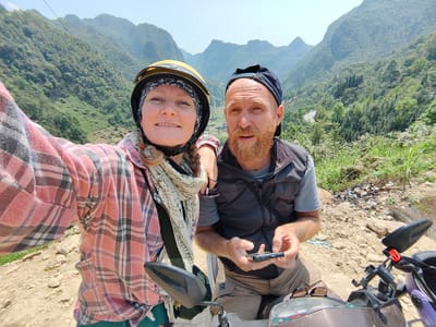 Two people are posing for a selfie on a motorcycle, with a mountainous landscape in the background. The woman on the left is wearing a pink plaid shirt, a scarf, and a yellow helmet with a patterned head covering underneath. She is smiling and extending her arm to take the photo. The man on the right has a beard and is wearing a baseball cap and a dark vest over a gray t-shirt. He is looking at the camera while holding a smartphone. They are seated on a motorcycle, with the handlebars and side mirrors visible in the foreground. The backdrop features rolling hills covered in green vegetation, a winding road, and a bright, clear sky. The location is Phố Bảng Commune, Vietnam. The foreground terrain is rocky and uneven. There is also trash on the ground near the man.