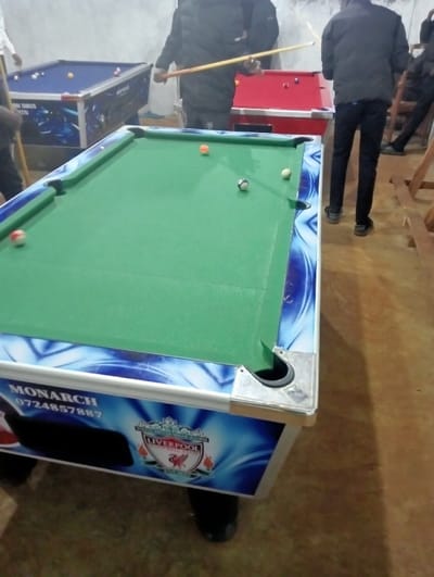 A billiards table with a green surface dominates the foreground, featuring balls scattered across it. The table's base displays a blue and white design, branded with "MONARCH" and a Liverpool FC logo. Behind it, a red and a blue billiards table are partially visible, each with balls on their surfaces. People are seen playing, with one holding a cue stick. The floor is light brown, and the overall lighting appears dim.