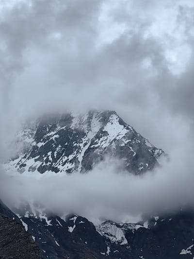 The image depicts a majestic mountain peak partially obscured by thick, swirling clouds. The mountain is rugged and imposing, with a dramatic contrast between its dark, rocky slopes and the white snow that clings to its upper reaches. The snow appears to be unevenly distributed, suggesting steep inclines and crevices.
The sky is overcast with heavy, grey clouds that create a dramatic and moody atmosphere. These clouds are not uniform; some are denser and darker, while others are lighter and more diffused, giving the impression of wind and movement. The clouds seem to wrap around the mountain, partially hiding its summit and creating a sense of mystery and awe.
In the foreground, the lower slopes of another mountain range are visible. These slopes are dark and rocky, with patches of snow scattered across them. The texture of these rocks is rough and detailed, indicating a natural, untamed environment. The overall lighting is subdued, characteristic of a cloudy or overcast day, likely in the late afternoon or early morning, though the exact time is not discernible.
The scene conveys a sense of raw, natural beauty and the power of nature. The clouds lend an ethereal quality to the mountain, making it appear almost mystical. The location appears to be a high-altitude mountainous region, possibly in the Himalayas, given the context of Gemur, India. There is no visible text in the image. The image captures a moment of dramatic weather interacting with a formidable landscape, evoking feelings of insignificance in the face of nature's grandeur.
