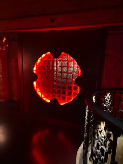 The image depicts a dimly lit interior with a prominent red glow emanating from a uniquely shaped window or illuminated feature in the center. This feature is textured with a grid pattern, resembling glass blocks. To the right, a dark, ornate railing suggests a staircase. The overall ambiance is dark, with red light casting shadows and reflections on the floor and walls. The location is specified as Khamkhi, Russia. No people or specific actions are visible.