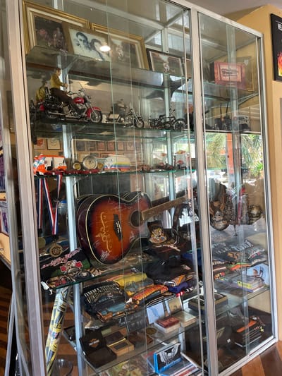 This image, captured in Ban Yid, Thailand, displays a large, multi-shelved glass display cabinet filled with an assortment of memorabilia and merchandise. The cabinet features silver-colored framing and clear glass panels, exhibiting reflections of an outdoor scene with green foliage and bright light.

The top shelf contains framed photographs, including a black-and-white portrait of two individuals in the center. Model motorcycles are prominently featured; on the left, a red cruiser-style motorcycle has two action figures, one driving with a yellow shirt and brown pants, and another seated behind. Several smaller model motorcycles and a vintage-style bicycle model are positioned to the right.

The second shelf from the top showcases various small vehicle models, including a white and orange tuk-tuk-like vehicle and a rainbow-striped Volkswagen van model. A clock with two circular gauges and small red model cars are also present. A large acoustic guitar with a sunburst finish lies horizontally, displaying numerous handwritten signatures on its body. To the left, a red, white, and blue ribbon with an attached medal hangs.

The third shelf from the top is stocked with folded T-shirts, many featuring designs such as "COMMANDER THAILAND BURIRAM 2019" and other motorcycle-related themes. A black and white patterned flag or banner is rolled and leaning on the far left. A dark, decorative, possibly metallic sculpture, which appears to be a motorcycle engine or part, is visible on the right.

The bottom shelf contains various items, including books, one explicitly titled "SPORTSTER MODELS" with a Harley-Davidson eagle logo. Several small boxes, one black with a "HARLEY-DAVIDSON MOTOR CYCLES" logo, and a golden disc are also present. More folded T-shirts and additional books, including one marked "2011," fill the remainder of the shelf.

The cabinet is positioned against a light yellow wall, with a wooden floor visible in the foreground. A partial black sign with red flame imagery and white text is visible on the wall to the far right of the cabinet.