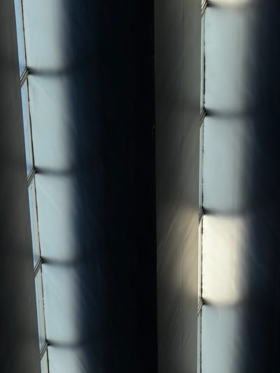 The image depicts an interior architectural scene, devoid of human presence or activity. A vertical assembly of six translucent rectangular panels, likely window panes or louvers, runs along the left edge of the frame. These panels are separated by narrow, dark horizontal bars and framed by a light-colored vertical structure. Natural light, entering from the upper-left, illuminates these panels and casts sharp, elongated rectangular shadows onto a contiguous, light-toned wall or ceiling surface. The shadows create a repeating pattern of dark, horizontally oriented rectangular bands against the brighter background. The central and upper-right area of the image is dominated by a deep shadow, rendering surfaces uniformly dark. A vertical seam or structural edge is faintly visible along the right side, with partial illumination revealing some surface texture. The surfaces appear generally smooth, with minor variations and potential dust or debris on the window frame. The specific location within Veracruz, Mexico, cannot be identified based solely on the visual information present.