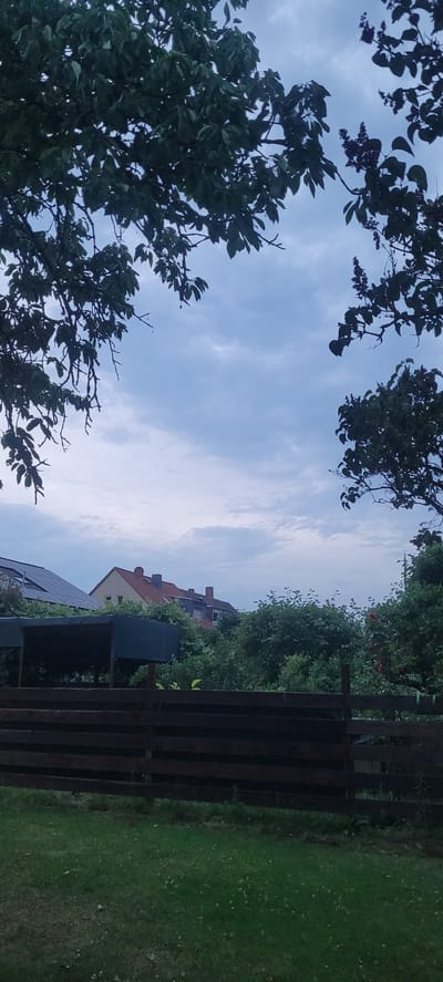 The image depicts a view of residential buildings and a garden in Dietzenbach, Germany, framed by tree branches. In the foreground, a dark wooden fence separates a patch of green grass from the rest of the scene. Behind the fence, there is dense green foliage and trees.

In the mid-ground, several houses are visible. One house, with a red-tiled roof, has visible windows and a satellite dish on its roof. To its left, another building appears to have solar panels installed on its roof. A covered structure, possibly a carport or shelter, is positioned in front of the buildings.

The background consists of a cloudy, overcast sky in shades of blue and grey. The overall impression is that of a suburban residential area on a cloudy day. No people are present in the image.