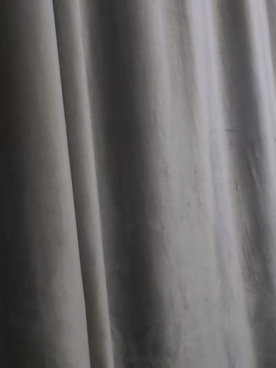 The image shows a close-up of a light-colored, draped fabric, likely a curtain or textile. The fabric exhibits soft folds and creases, creating a play of light and shadow. The texture appears smooth with a subtle sheen. There are a few small, dark smudges on the fabric, suggesting a potential need for cleaning. The overall impression is of a simple, domestic textile. The image does not contain any specific elements that would allow for its location to be identified within the city of Жарлыкөл, Kazakhstan, or any other urban environment.