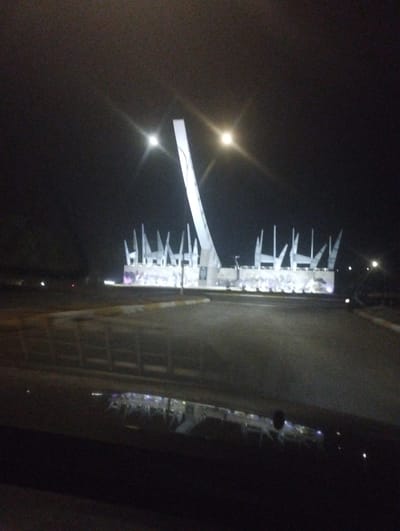 The image shows a nighttime view of a monument in Campo Carabobo, Venezuela. The monument features a tall, curved white tower and a lower section with several vertical white structures resembling spikes or spears. The monument is illuminated, creating a bright contrast against the dark sky. The background is mostly dark, with faint lights suggesting other buildings or infrastructure in the distance. The foreground shows a paved road, with reflections visible on the surface. The photo appears to be taken from inside a vehicle, as part of the dashboard is visible at the bottom of the frame.