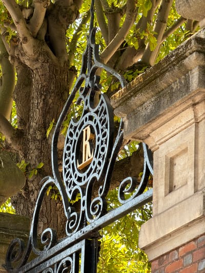 The image shows an ornate black iron gate with an ornamental emblem featuring the letter "R" in gold. The gate is part of a larger structure, with a stone pillar to the right and a large tree with textured bark and green leaves behind the gate. The stone pillar is made of light-colored, rough-textured blocks with some darker accents and has a decorative cornice. The tree has thick, gnarled branches. Sunlight filters through the leaves, creating bright highlights and dappled shadows. The scene appears to be outdoors, possibly an entrance to a park or an estate. The location is the city of Courbevoie, France.