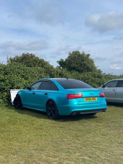 The image shows a turquoise Audi A6 parked on a grassy field with green foliage and trees in the background, under a partly cloudy sky. The car's license plate reads "OV64 HSE". Another vehicle, partially visible, is parked nearby. The location is possibly in Sevenoaks, United Kingdom, though no definitive landmarks are visible. There are no people visible in the image.