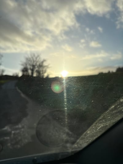 The image presents a rural landscape scene at low sun, viewed through a dirty and scratched vehicle windshield in Shelve, United Kingdom. The sun, positioned near the horizon slightly right of center, emits prominent lens flares, including a distinct green circular flare and vertical light streaks. A paved pathway or road curves from the lower-left foreground into the mid-ground, flanked by green grass and terrain. A cluster of leafless deciduous trees is silhouetted against a partially cloudy sky on the left horizon, with additional distant foliage or terrain visible on the right. The vehicle's interior, possibly a circular vent or dashboard element, is faintly reflected in the lower-center of the windshield. No people, animals, or specific interactions are present in the visible outdoor environment.