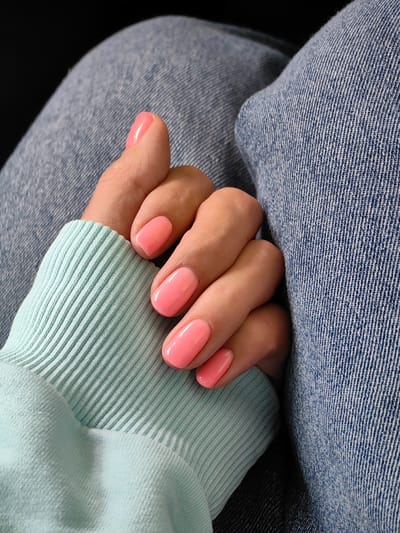 The image shows a close-up view of a person's left hand, which is resting on light blue denim jeans and a mint green sweater. The fingernails are painted a bright, glossy pink. The hand is positioned so that the thumb and index finger are visible, along with parts of the middle and ring fingers. The focus is on the nails and the textures of the clothing. There is no discernible location in the city of Фаніпальскі сельскі Савет, Belarus visible in this image.