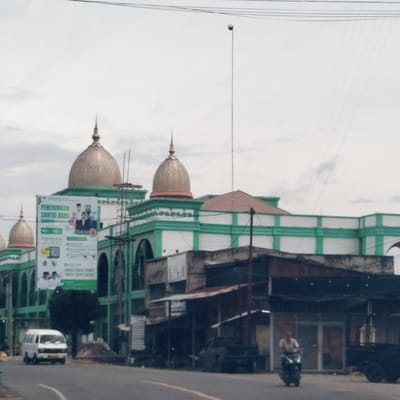 This image captures a street scene in Keudejunieb, Indonesia, featuring a prominent building with golden domes and green and white facades, likely an Islamic boarding school or mosque. A large banner hangs from this building, advertising "PENERIMAAN SANTRI BARU" (New Santri Admission) along with contact phone numbers (0822 1002 6180 and 0822 3247 2243), a QR code, and images of people and the number "10 10".
In the foreground to the right, there are several smaller, older buildings or stalls with corrugated metal roofs. A person is seen riding a motorcycle on the street in front of these structures, moving towards the right. On the left side of the road, a white van is parked or driving. The road itself has a faint white curved line and a partial yellow line. A small tree is visible between the van and the main building.
The sky is overcast, suggesting a cloudy day with soft, diffused lighting. Overhead, a tall, slender pole with a spherical top rises high into the sky, along with multiple thin utility lines stretching across the upper part of the frame. The overall atmosphere is that of a typical, functional street in a town or city.