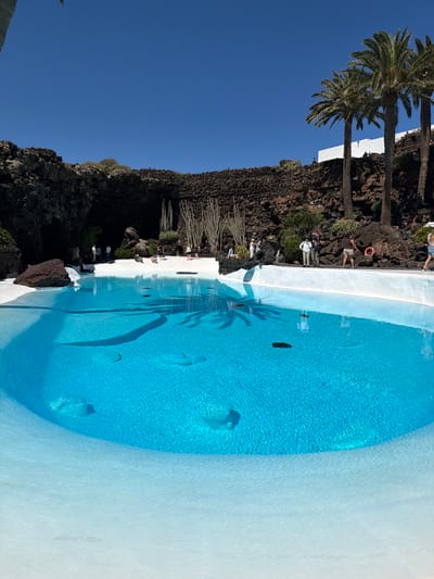 This image captures a beautifully designed swimming pool set within a dramatic volcanic landscape under a clear blue sky. The pool features bright blue water with visible ripples and reflections, including the prominent shadow of a palm tree. Its edges are defined by smooth, pristine white concrete, which contrasts sharply with the dark, rugged volcanic rock formations that surround it.

Several palm trees with green fronds stand tall, particularly on the left side of the frame, alongside some cactus-like plants growing among the rocks. Numerous individuals are visible around the pool and on the surrounding rock pathways. They appear to be visitors or tourists, some standing and observing, while others are seated, enjoying the unique environment. One person is seen near a red lifebuoy.

The bright, cloudless sky and strong shadows indicate a clear, sunny day. The scene evokes a sense of tranquility and awe, highlighting the harmonious integration of man-made structures with the natural, volcanic terrain. This aesthetic, combining white architecture with dark rock and lush plants, is characteristic of attractions in Lanzarote, Spain, such as Jameos del Agua, which is consistent with the provided location context of Haría. No visible text is present in the image.