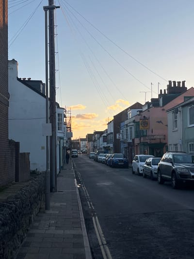This image displays a narrow urban street in Weymouth, United Kingdom, under low sunlight, suggesting late afternoon or early evening. The sky is predominantly clear blue with scattered clouds near the horizon, illuminated by warm, golden hues, casting the street and buildings into shadow.
On the left, a low stone wall borders a grey paved sidewalk. A tall wooden utility pole, supporting multiple cross arms and numerous radiating wires, stands prominently. A second, thinner pole is adjacent to it. Further down the sidewalk, one pedestrian walks away from the viewer. Buildings lining the left side include a white-rendered structure with a bay window; scaffolding is visible on a building further down the street.
The asphalt road surface displays continuous double yellow lines. A continuous line of various cars and SUVs is parked bumper-to-bumper along the right street edge. Buildings on the right feature varied facades, including a pink structure housing "The Dolphin" pub, identifiable by its circular sign depicting a crest and a dolphin mural on the wall to its right. Rooftops are visible against the sky, featuring chimney pots and antennas. Vehicles are visible in the distant street.