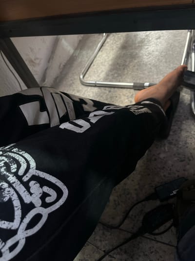 A low-angle shot shows a person's legs from the knee down, draped over metal chair legs beneath a dark wooden surface. The person is wearing black pants with large white graphic designs. A bare foot is visible, resting on a black object, possibly a phone. Electrical cords and a black charging adapter are on the speckled concrete floor. The background is out of focus, showing a textured wall and more chair legs. The scene appears to be indoors, possibly in Alagbede, Nigeria, with evidence of personal belongings and electronic devices.