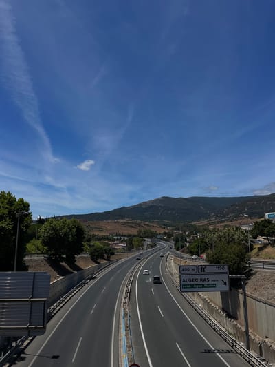 The image shows a highway on a sunny day, with a clear blue sky and some wispy clouds. The highway curves to the right, with several lanes of traffic. Cars are visible on the road.

On the left side of the highway, there is a steep, tree-covered hillside. In the distance, a mountain range is visible. A town or village is nestled at the base of the hill.

A white highway sign is mounted on a pole beside the road. The sign has two panels. The top panel is black with white text and a white arrow pointing to the right, indicating "ALGECIRAS oeste" and "1120". Below this, a smaller white panel with black text reads "400 m". The bottom panel of the sign is green with white text and a white arrow pointing to the right, indicating "Avda. Agua Marina".

The sun appears to be high in the sky, suggesting it is midday. The weather is clear and warm. The scene is likely a major road leading into or out of Algeciras, Spain, as indicated by the signage. There are no people clearly visible in the image. The overall impression is one of travel and transit through a scenic, possibly Mediterranean landscape.