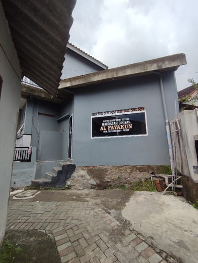 The image shows the exterior of a building in the city of Kecamatan Prigen, Indonesia (latitude: -7.675, longitude: 112.66). The building has a grey painted wall, a roof made of corrugated metal, and a sign that reads “KANTOR SEKRATARAT YAYASAN MADRASAH DINIYAH AL FAYAKUN” with the address “DS. CAMBIRAN - PRIGEN”. In front of the building, there is a set of concrete steps leading to a doorway that is partly obscured by a low wall. The ground in front of the building is paved with brick tiles. The building is situated between two other structures, with the roof of one of them partially visible in the foreground. The sky is overcast with grey clouds.