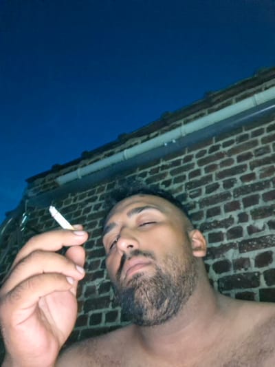 This is a slightly angled, close-up, low-light photograph of a person lying down, seen from the chest up. The person has dark skin, a beard, and appears to be holding a lit cigarette in their right hand, near their face. Their eyes are closed, and their mouth is slightly open. They are lying against a brick wall, which is visible in the background. The sky is a deep blue, suggesting it is either dawn or dusk. A white downspout runs vertically along the brick wall. The overall mood of the image is relaxed, perhaps even contemplative. The lighting suggests an outdoor setting at either dawn or dusk. The location context is Evere, Belgium. There is no visible text in the image.