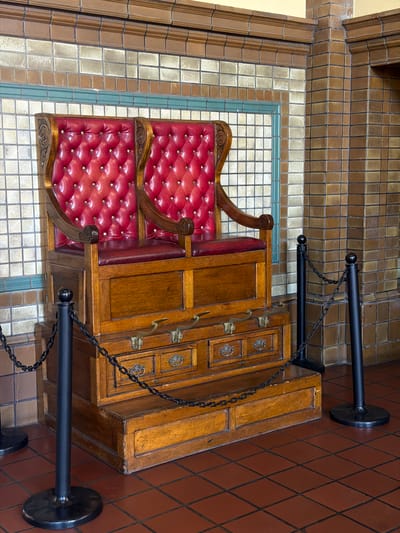 An unoccupied, vintage double shoe shine stand is centrally positioned within an interior tiled room, identified as the Santa Fe Depot in San Bernardino, United States of America. The stand is constructed from polished light-brown wood, featuring two elevated seats. Each seat has a tufted red leather backrest with brass buttons and a solid red leather seat cushion. The armrests are crafted from carved wood. The wooden base incorporates two smaller drawers with ornate brass pulls, two larger drawers with brass latches, and two brass-colored footrests. A wooden step extends from the base for access. The walls are clad in ceramic tiles, featuring a pattern of beige and light-brown square tiles in the lower section, separated by a horizontal green tile band from larger brown rectangular tiles above. A decorative brown tiled molding runs along the upper perimeter. The floor consists of reddish-brown square ceramic tiles with dark grout. Two black stanchions with spherical tops are linked by a black metal chain, forming a barrier in front of the stand. No individuals are present, and no actions or interactions are depicted.